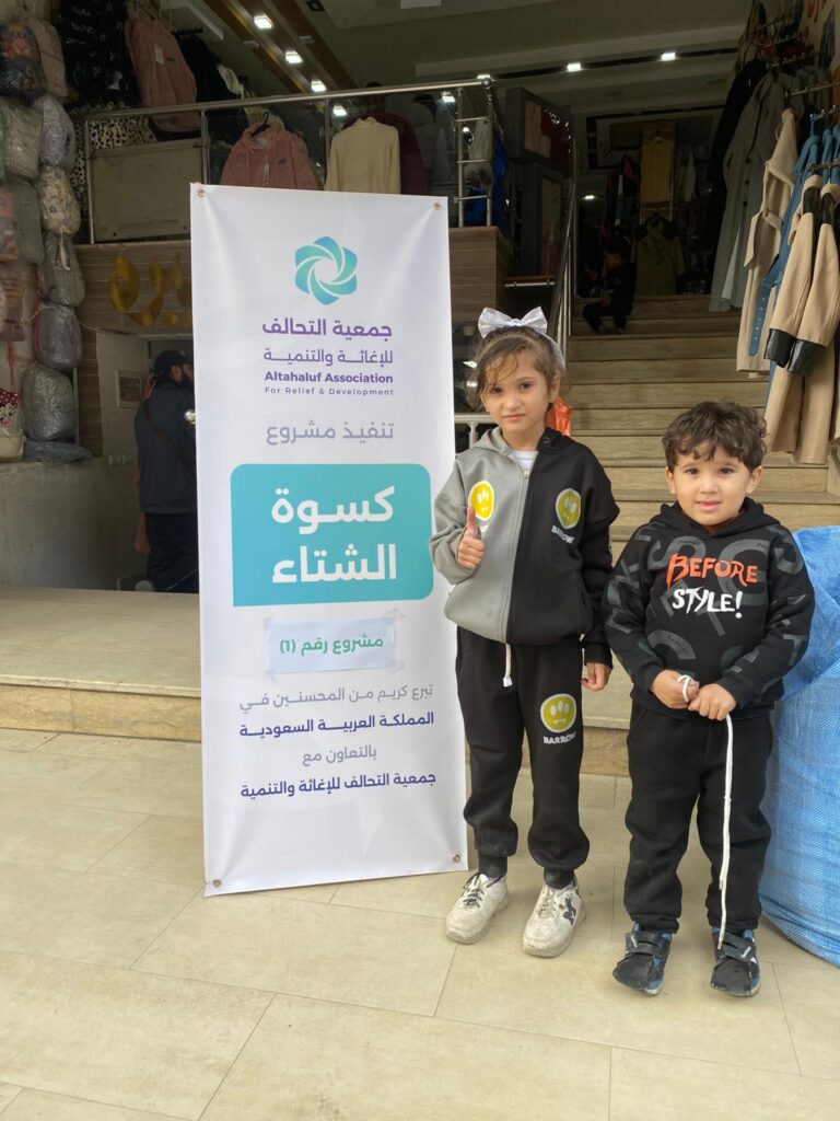 أطفال غزة أمام لافتة مشروع كسوة الشتاء لجمعية التحالف للإغاثة والتنمية